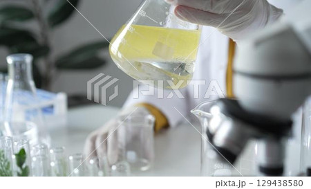 A scientist wearing white protective gloves is shaking up a yellow chemical solution inside an Erlenmeyer flask in a laboratory set, close up. Science and medicine concepts A scientist wearing white protective gloves is shaking up a yellow chemical solution inside an Erlenmeyer flask in a laboratory set, close up. Science and medicine concepts 129438580