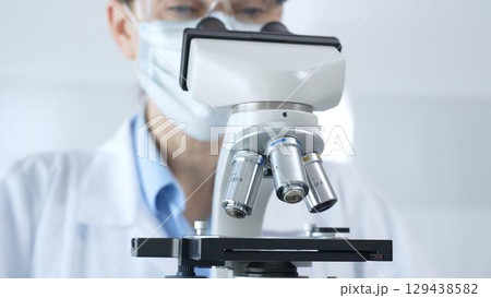 Scientist wearing lab coat and protective glasses using a modern microscope in a laboratory setting, conducting research and experiments in a scientific environment. Health care and science concept 129438582