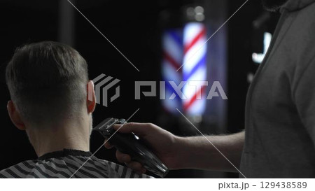 Male professional barber trimming client's hair with electric clipper, vintage illuminated barber pole visible in background, creating contemporary grooming experience Male professional barber trimming client's hair with electric clipper, vintage illuminated barber pole visible in background, creating contemporary grooming experience 129438589