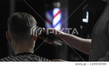 Male professional barber trimming client's hair with electric clipper, vintage illuminated barber pole visible in background, creating contemporary grooming experience 129438590