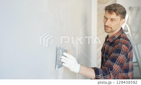 Construction worker man wearing gloves and holding a putty knife is applying plaster on a white wall, performing home renovation and improvement Construction worker man wearing gloves and holding a putty knife is applying plaster on a white wall, performing home renovation and improvement 129438592