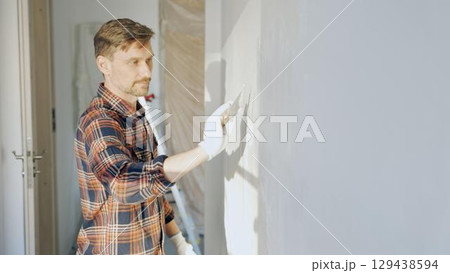 Male construction worker wearing protective gloves smoothing plaster onto wall surface during home renovation using professional trowel tool 129438594