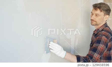 Construction worker man wearing gloves and holding a putty knife is applying plaster on a white wall, performing home renovation and improvement Construction worker man wearing gloves and holding a putty knife is applying plaster on a white wall, performing home renovation and improvement 129438596