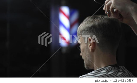 Professional barber cutting hair of male client in barbershop, using scissors and comb, with barber pole in background, creating stylish and modern haircut 129438597