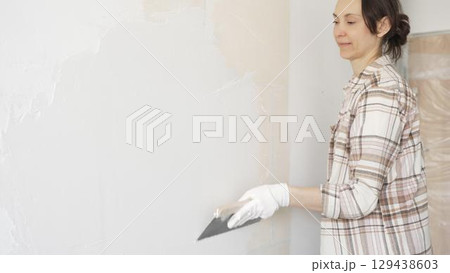 Woman applying plaster to wall with putty knife, wearing protective gloves and creating smooth surface during home renovation work 129438603