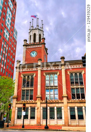 Under dramatic clouds, the red brick Manchester City Hall stands on Elm Street in Manchester, New Hampshire, United States, its clock tower and Gothic windows framed by spring greenery 129439229