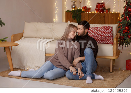 Loving young couple sitting on floor near sofa in cozy room with Christmas tree and lights Loving young couple sitting on floor near sofa in cozy room with Christmas tree and lights 129439895