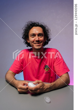 Vertical portrait of cheerful young adult man with curly hair and pink t-shirt cutting fingernails using nail clipper sitting at gray table against purple background, smiling looking at camera. Vertical portrait of cheerful young adult man with curly hair and pink t-shirt cutting fingernails using nail clipper sitting at gray table against purple background, smiling looking at camera. 129440096