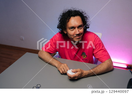 Portrait of joyful young man with curly hair and pink t-shirt cutting fingernails using electric nail clipper sitting at gray table against purple background, smiling looking at camera. Portrait of joyful young man with curly hair and pink t-shirt cutting fingernails using electric nail clipper sitting at gray table against purple background, smiling looking at camera. 129440097