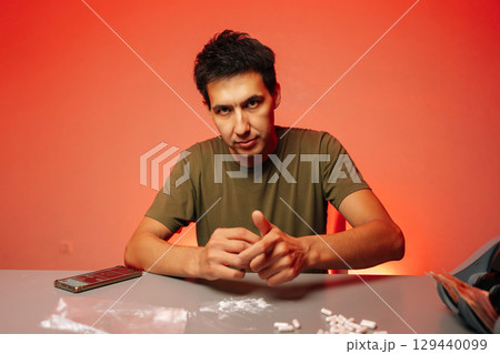Portrait of serious drug realer male preparing dose of cocaine or heroin, surrounded by pills, cash and smartphone on red background, highlighting dangers of addiction and illegal drug trade. 129440099