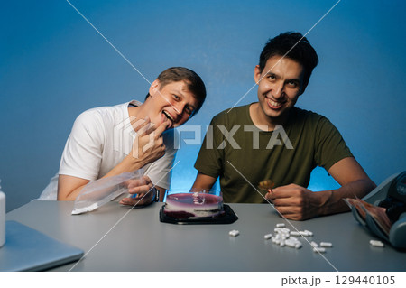Two cheerful friends sitting at table with drugs, white powder, pills, cake, money in studio with isolated blue background. Concept of party atmosphere with substance abuse. Two cheerful friends sitting at table with drugs, white powder, pills, cake, money in studio with isolated blue background. Concept of party atmosphere with substance abuse. 129440105