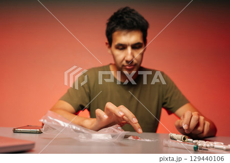Selective focus shot of drug realer male preparing dose of cocaine or heroin, surrounded by pills, cash and smartphone on red background, highlighting dangers of addiction and illegal drug trade. 129440106
