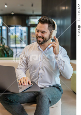 Smiling young businessman using laptop and listening to music via headphones in modern office lobby 129440422