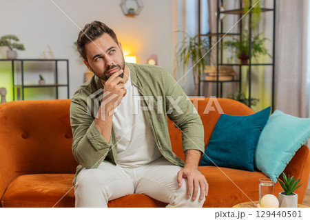 Sad lonely Caucasian young man sitting on sofa at home room looks pensive thinks over life concerns 129440501