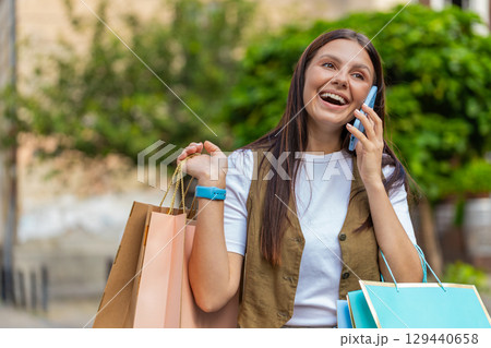 Happy young woman shopaholic carrying shopping bags while talking via smartphone on city street Happy young woman shopaholic carrying shopping bags while talking via smartphone on city street 129440658