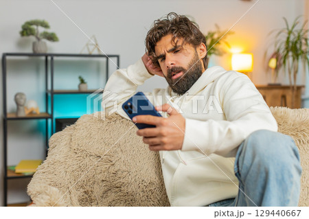 Upset displeased Indian man guy using smartphone reading bad news sitting on couch in living room 129440667