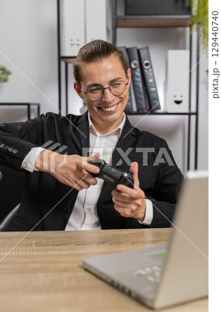 Excited young businessman playing video game on laptop with joystick controller at home office desk 129440740