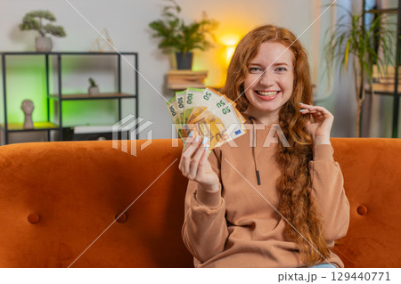 Successful rich young Caucasian woman holding euro bill money fan sitting on sofa couch at home Successful rich young Caucasian woman holding euro bill money fan sitting on sofa couch at home 129440771
