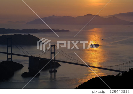 愛媛県　今治市　夕日に染まる海で本州と四国を結ぶ来島海峡大橋に近づく船 129443832