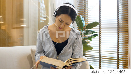 Happy asian woman use headphones listen to music reading book sitting on cozy sofa in living room at home. Female asian people work at home research book read magazine smiling happy reader lifestyle Happy asian woman use headphones listen to music reading book sitting on cozy sofa in living room at home. Female asian people work at home research book read magazine smiling happy reader lifestyle 129444684