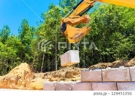 Heavy machinery is lifting concrete block at construction site surrounded by trees under build retaining walls 129451350