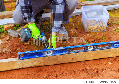 Contractor in gloves drills into wood while using level to ensure accuracy in backyard construction project. Contractor in gloves drills into wood while using level to ensure accuracy in backyard construction project. 129451459