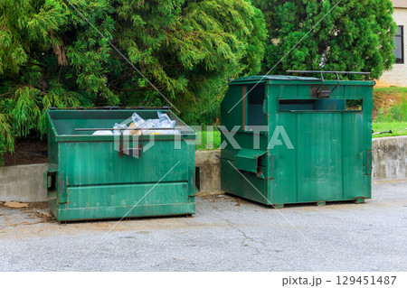 Two green dumpsters are located next to concrete wall, filled with recyclable materials, under brighter sunlight. 129451487