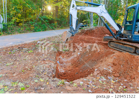 Heavy machinery digs into earth, clearing space near road surrounded by trees in during construction work Heavy machinery digs into earth, clearing space near road surrounded by trees in during construction work 129451522