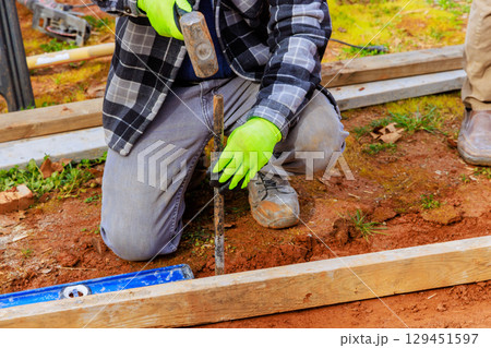 Contractor worker kneels on ground with hammer, driving stake into soil while preparing for construction. 129451597