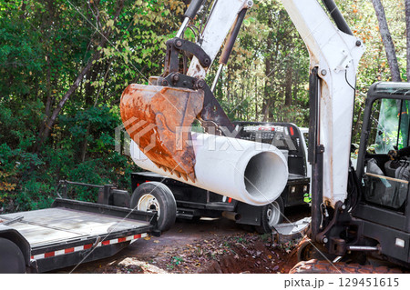 Construction workers use an excavator to lift concrete drainage pipe from truck for utility installation in forest. Construction workers use an excavator to lift concrete drainage pipe from truck for utility installation in forest. 129451615