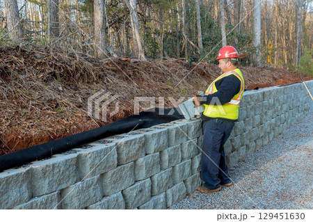 Worker arranges stone blocks for retaining wall by gravel road amid trees. 129451630