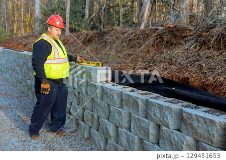 Worker in safety gear levelling measures wall blocks to ensure proper alignment along gravel path 129451653