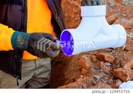 Construction worker uses purple primer on PVC pipe fitting at job site, preparing for plumbing installation sewage pipes work. 129451710