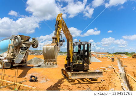 Excavator lifts dirt while concrete mixer pours materials at construction site under works day Excavator lifts dirt while concrete mixer pours materials at construction site under works day 129451765