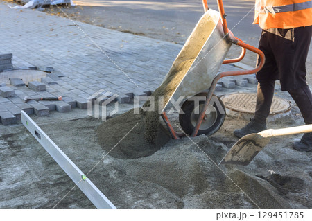 Construction worker pours sand from wheelbarrow onto ground to prepare for paving bricks during works day. Construction worker pours sand from wheelbarrow onto ground to prepare for paving bricks during works day. 129451785