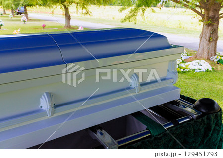 White casket is positioned in cemetery during funeral service among green trees floral tributes. 129451793