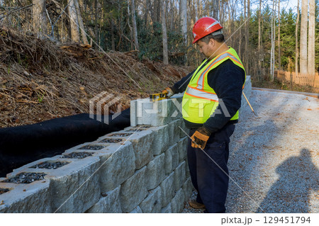 Construction worker measures stone blocks while working on retaining wall beside gravel road Construction worker measures stone blocks while working on retaining wall beside gravel road 129451794