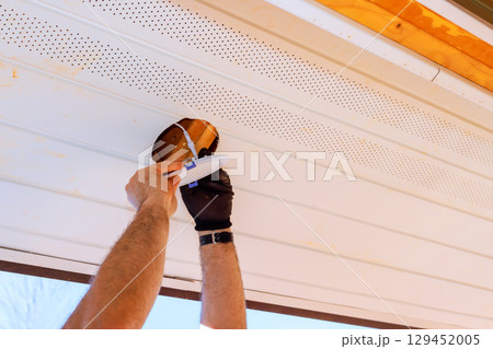 Homeowner applies lighting fixture in ceiling in soffit under eaves of house in suburb. 129452005