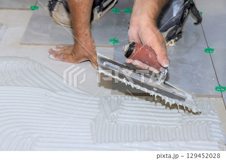 Worker uses trowel to spread adhesive while laying tiles on construction site in broad work day 129452028