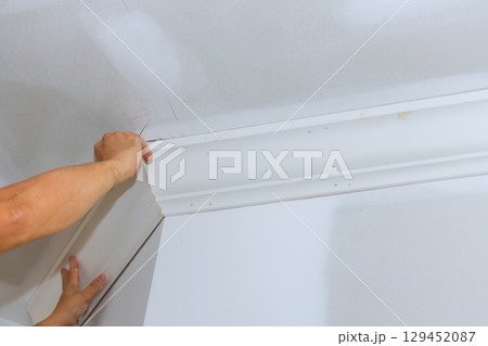 Carpenter carefully aligns, installs crown molding at corner of white room to enhance its appearance design. 129452087