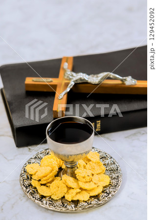 Silver cup filled with wine plate of wafers placed by Bible and crucifix during morning prayer. 129452092