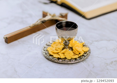 Chalice filled with wine plate of wafers are arranged on marble table with cross an open Bible book. 129452093
