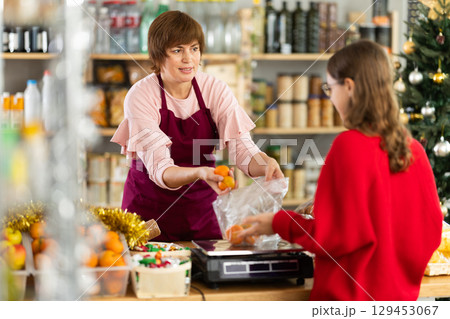 Woman sells fruits to teen girl, puts tangerines in bag, puts them on scale and weighs fruits Woman sells fruits to teen girl, puts tangerines in bag, puts them on scale and weighs fruits 129453067