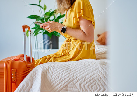 Woman in dress sitting on bed with orange suitcase using smartphone indoors Woman in dress sitting on bed with orange suitcase using smartphone indoors 129453117
