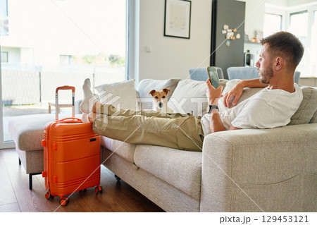 Man sitting on sofa with suitcase and using smartphone 129453121