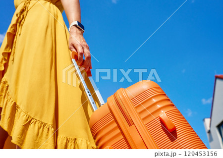 Woman in yellow dress holding orange suitcase under blue sky Woman in yellow dress holding orange suitcase under blue sky 129453156