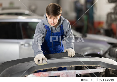 Young auto mechanic polishes car bumper before painting. Recovery and repair after an accident Young auto mechanic polishes car bumper before painting. Recovery and repair after an accident 129453546