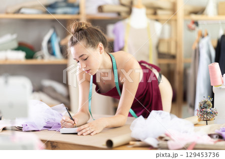 Seamstress in black stockings and an apron draws the outline of dress using patterns Seamstress in black stockings and an apron draws the outline of dress using patterns 129453736