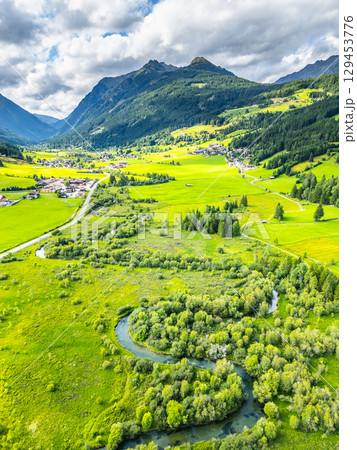 A winding river flows gracefully through a vibrant green valley surrounded by majestic mountains in the Alps. The landscape is dotted with patches of trees and meadows, showcasing nature's beauty. 129453776