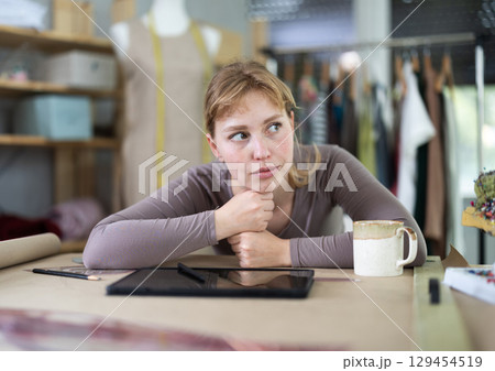 Thoughtful female fashion designer sitting at work table in studio 129454519